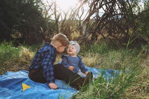 A little boy in a plaid shirt sits on a blue tarp and gently leans toward a smiling baby in a sunlit grassy woodland.