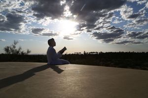 Person in white sits on a flat rock holding an open book, silhouetted against a glowing sun and dramatic cloudy sky.