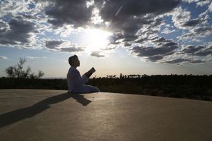 Silhouetted person sits on a flat rock reading a book as the sun breaks through dramatic clouds over the desert.
