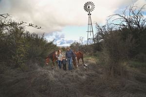 A family with a young child walks along a grassy trail with two horses and a dog near a windmill under a cloudy sky.