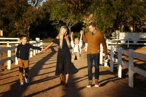 Parents swing a toddler between them while walking with their older son along a white-fenced path lined with trees.