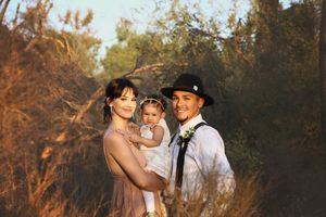 A young family of three stands among golden desert brush at sunset, with parents holding their smiling baby girl.