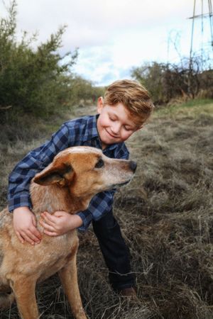 A smiling boy in a blue plaid shirt hugs a tan ranch dog in a grassy field with shrubs and a cloudy sky behind.