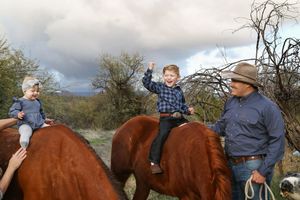 A toddler and a young boy sit on chestnut horses while a man in a cowboy hat stands beside them outdoors.