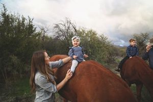 A woman steadies a smiling toddler sitting on a chestnut horse while a boy rides another horse nearby.