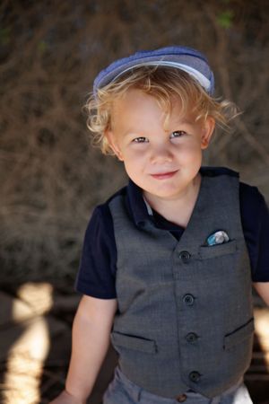 Smiling toddler boy with curly blond hair wears a blue newsboy cap, gray vest and navy shirt while posing outdoors.