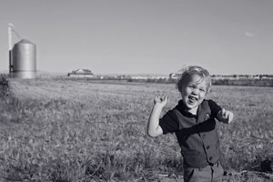 Black and white photo of a laughing little boy with arms raised in a grassy field with a grain silo in the distance.