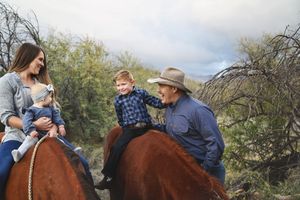 A smiling woman holding a toddler on horseback looks at a boy and a man in a cowboy hat on another horse.