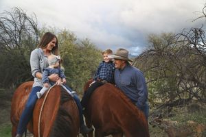 A woman with a toddler and a boy on separate horses ride beside a man in a cowboy hat through desert brush.