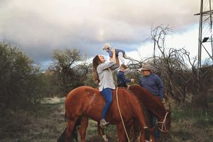 A woman on horseback lifts a laughing toddler overhead while a man in a cowboy hat and a boy ride nearby.