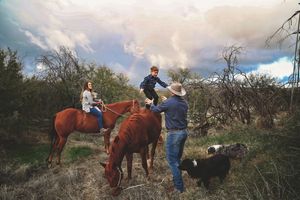 A woman on horseback smiles as a man in a cowboy hat helps a boy climb up while another horse grazes and a dog waits nearby.