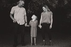 Black and white photo of a smiling couple holding hands with their young daughter, who looks up at them in a wooded setting.