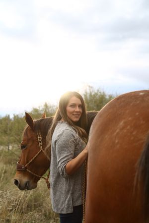 A smiling woman with long brown hair stands beside a chestnut horse in a field, lit by soft golden sunlight.