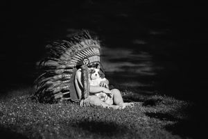 Black and white photo of a child in a feathered Native American-style headdress sitting on grass and holding a small dog.