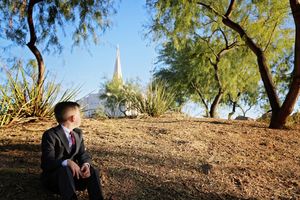 Boy in a dark suit and tie sits on a dry hillside, gazing at a distant white temple spire framed by desert trees and blue sky.