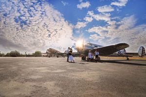Group of people stands beside vintage airplanes on a sunlit tarmac under a bright blue sky filled with scattered clouds.