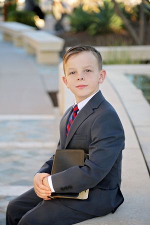 Young boy in a gray suit and striped tie sits beside a fountain, holding a black book and looking at the camera.