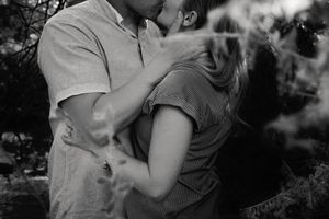 Black and white photo of a couple sharing a kiss outdoors, framed by soft blurred foliage in the foreground.