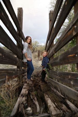 Smiling woman and young boy stand on an old weathered wooden bridge with rustic railings under a cloudy sky.