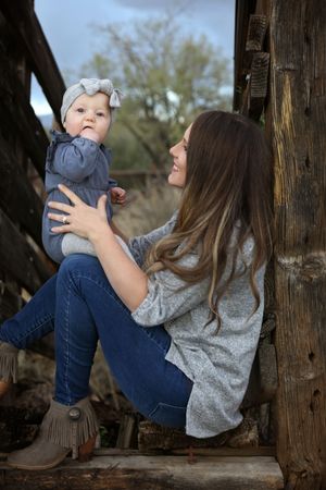 Smiling mother in jeans and a gray sweater sits on rustic wood holding her baby in a gray bow headband outdoors.
