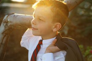 Boy in a white shirt and red striped tie holds his suit jacket over his shoulder, lit by warm golden sunlight.