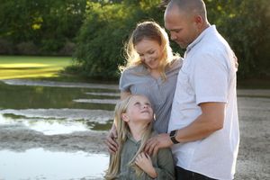 Smiling family of three embraces beside a pond, with the young blonde daughter looking up at her parents in soft evening light.