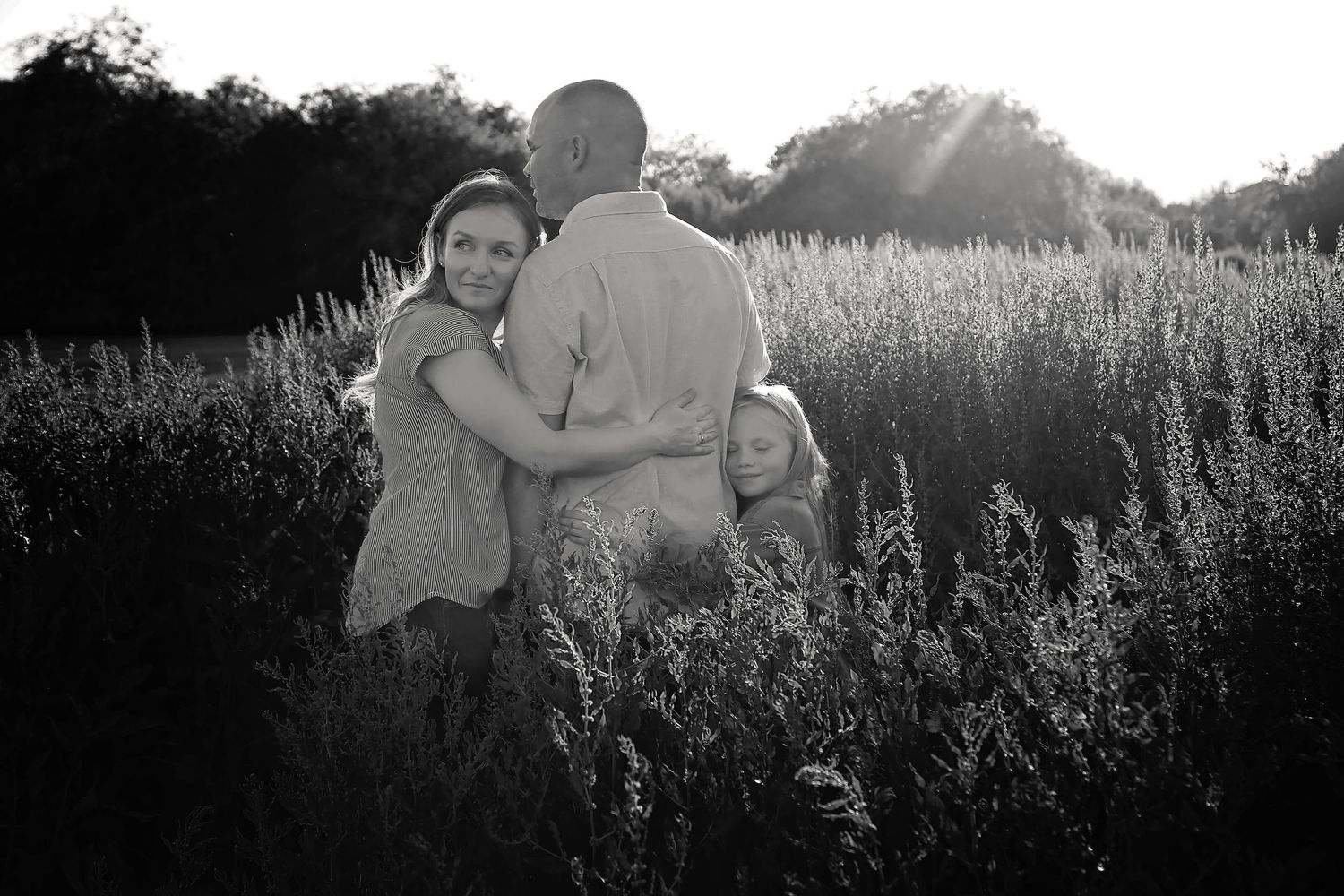 Black and white photo of parents hugging their young daughter in a tall grass field with sunlight streaming through the trees.