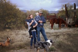 Family with a toddler stands outdoors on a ranch with a cowboy hat, two dogs, a brown horse, and a wooden fence behind them.