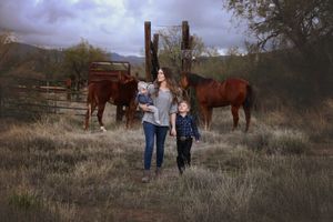 A mother holds a baby while walking with a young boy through a grassy ranch field with horses and a wooden fence behind them.
