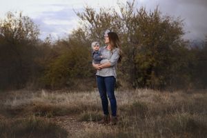 A mother in jeans and boots holds her baby while standing in an open field with trees under a moody evening sky.
