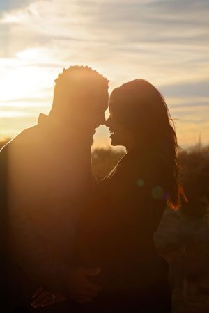 A couple stands forehead to forehead in silhouette against a glowing golden sunset sky in a romantic outdoor portrait.