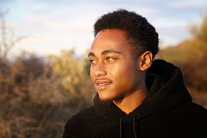 A young man in a black hooded sweatshirt gazes thoughtfully to the side in warm golden light with desert brush behind him.