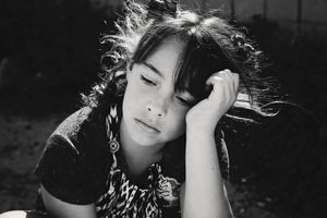 A black and white portrait of a young girl with wavy dark hair resting her head on her hand with a pensive expression.
