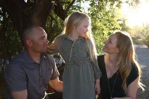 A father, mother and young daughter share a warm family moment outdoors beneath trees in soft golden sunlight.