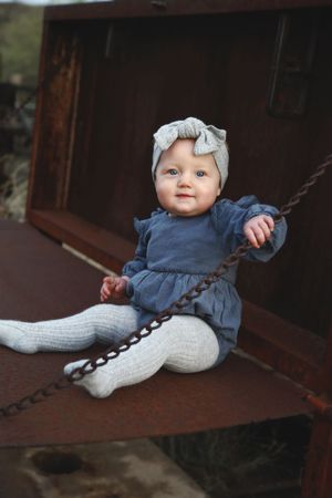 A smiling baby in a blue dress, gray bow headband and knit tights sits on a rusty metal surface beside a chain outdoors.