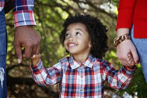 A smiling toddler in a plaid shirt holds the hands of two adults while standing outdoors among green trees.