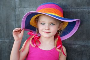 A smiling young girl in a colorful striped sun hat and pink tank top with pink ribbons poses against a gray wall.