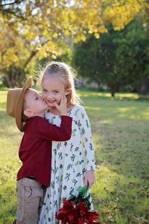 Little boy in a cowboy hat kisses a smiling girl in a floral dress on the cheek in a sunny autumn park.