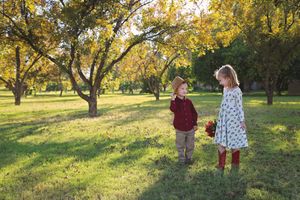 Young boy in a cowboy hat and girl in a floral dress holding red flowers stand in a grassy park under golden autumn trees.