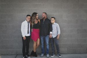 A family of four in stylish outfits, including two boys, a woman in a red skirt and a bearded man, pose together against a gray brick wall.