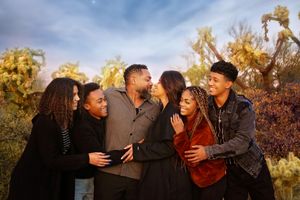 Smiling family of six embrace in a close group portrait among desert plants under a soft blue and golden sky.