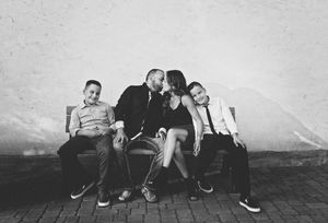 A black and white family portrait shows two boys sitting on a bench with their parents kissing between them against a textured wall.