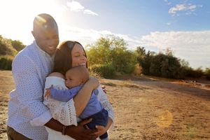 Smiling couple lovingly hug and hold their baby in a sunlit desert landscape with green trees in the background.