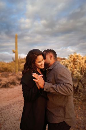 Romantic couple embrace and show an engagement ring at sunset with saguaro cactus and cloudy skies in the desert.