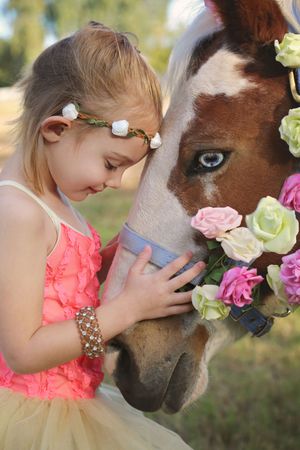Little girl in a pink dress and flower crown gently touches the face of a pinto horse adorned with pink and cream roses.