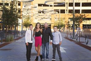 A family of four stands smiling on an urban plaza with modern hanging ring sculptures overhead, a mother, father and two boys.