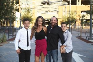 A smiling family poses together on a city plaza, with a mother in a pink skirt, a father in black, and two boys in dress shirts.