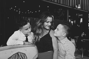 Black and white photo of a mother smiling at two boys in dress shirts as they lean toward her in a softly lit evening setting.
