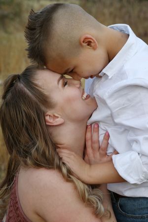 A smiling woman with long blonde hair touches foreheads with a young boy in a white shirt during an outdoor family portrait.