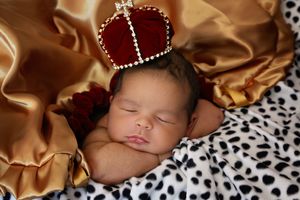 A sleeping newborn baby wearing a red velvet crown rests on gold satin and a black and white spotted blanket.